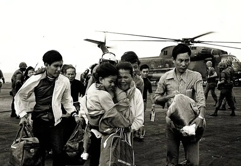 A group of people, some carrying bags and holding children, walk away from a military helicopter on a landing pad with armed soldiers in the background.