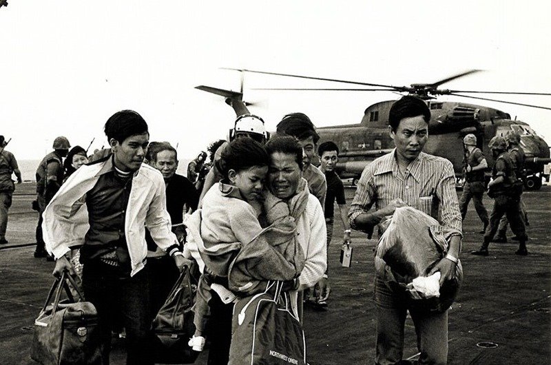 A group of people, some carrying bags and holding children, walk away from a military helicopter on a landing pad with armed soldiers in the background.