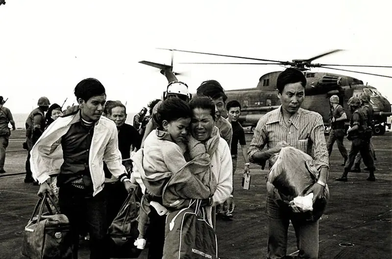 A group of people, some carrying bags and holding children, walk away from a military helicopter on a landing pad with armed soldiers in the background.